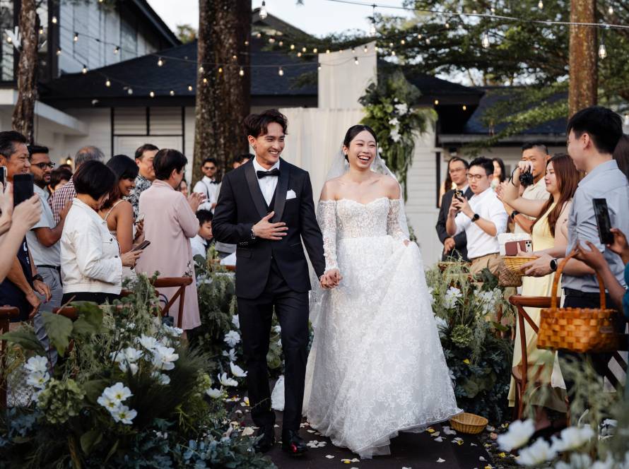 Bride in white lace gown and groom in black tuxedo walk down flower-lined aisle while guests clap and photograph at Petrichor Taiping garden wedding