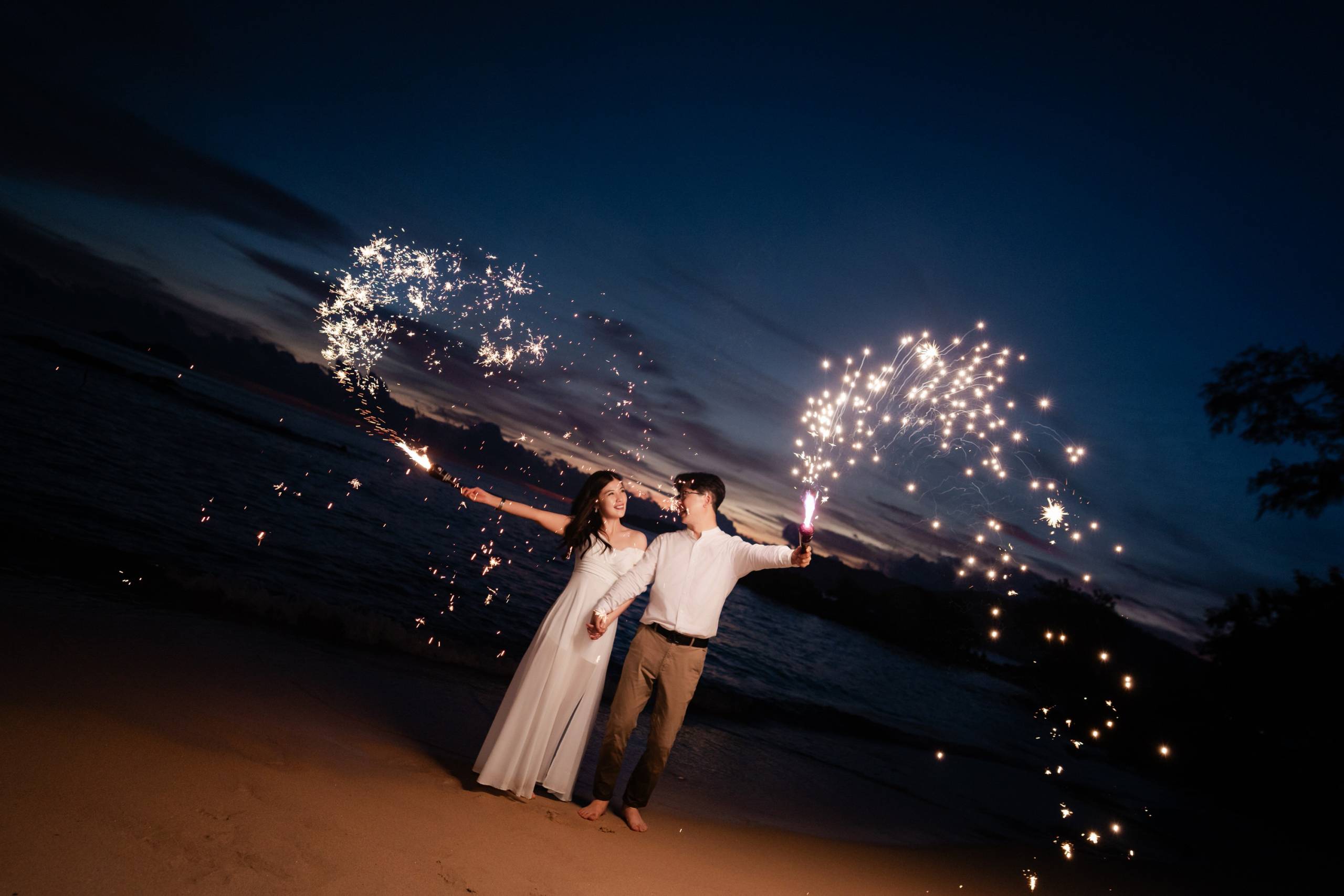 Groom in white shirt holds parasol over barefoot bride embraces his bride in quiet stillness along Teluk Bahang shoreline,...